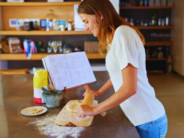 Cuisinière qui pétrit une pâte sur un plan de travail fariné, livre de recettes ouvert devant elle